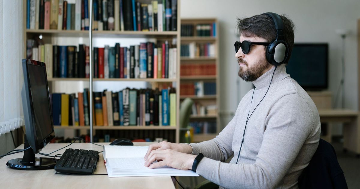Een blinde man met hoofdtelefoon werkt aan een computer met brailleleesregel