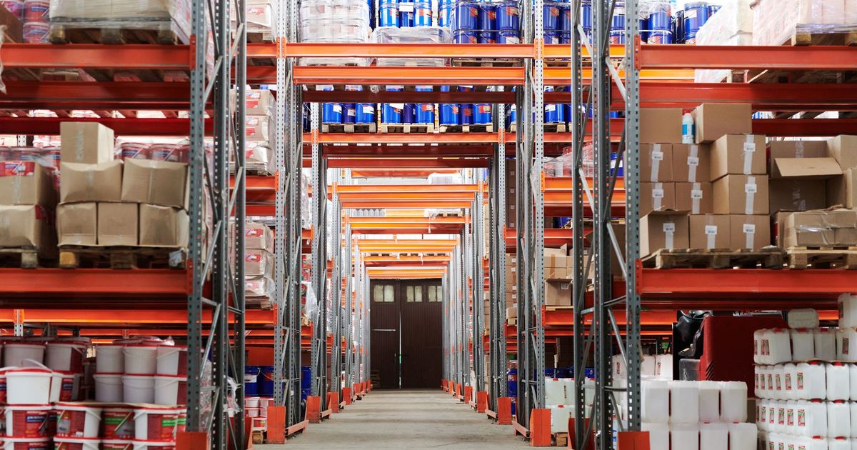 Een gangpad tussen hoge metalen palletrekken vol dozen, vaten en jerrycans in een magazijn